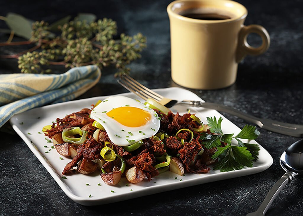 Harvest Shreds BBQ-spiced hash with new potatoes and leeks, and topped with a fried egg, on a square white plate with a fork. Folded yellow and blue napkin and a yellow mug of coffee.