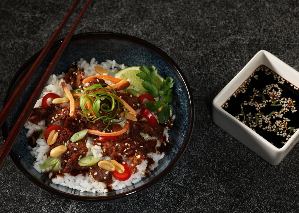Harvest Shreds spiced with Bulgogi Blend on a bed of rice, with red peppers, scallions, peanuts, carrots, and sesame seeds. In a large dark bowl with chopsticks and a square white ramekin with soy sauce and sesame seeds.