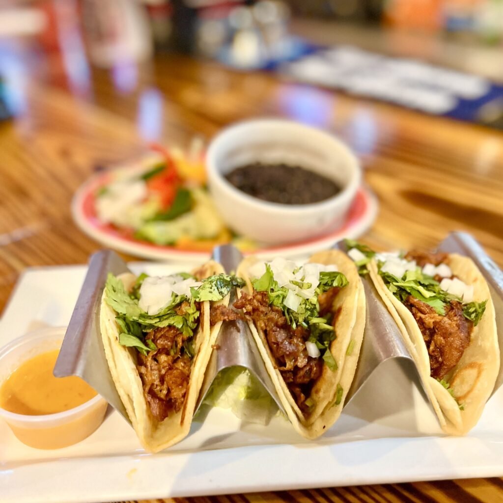 Three Harvest Shreds tacos in a taco holder, topped with salsa, cilantro and chopped white onions. Ramekin of dipping sauce, all on a square white plate. In the background, a bowl of black beans and fresh chilis