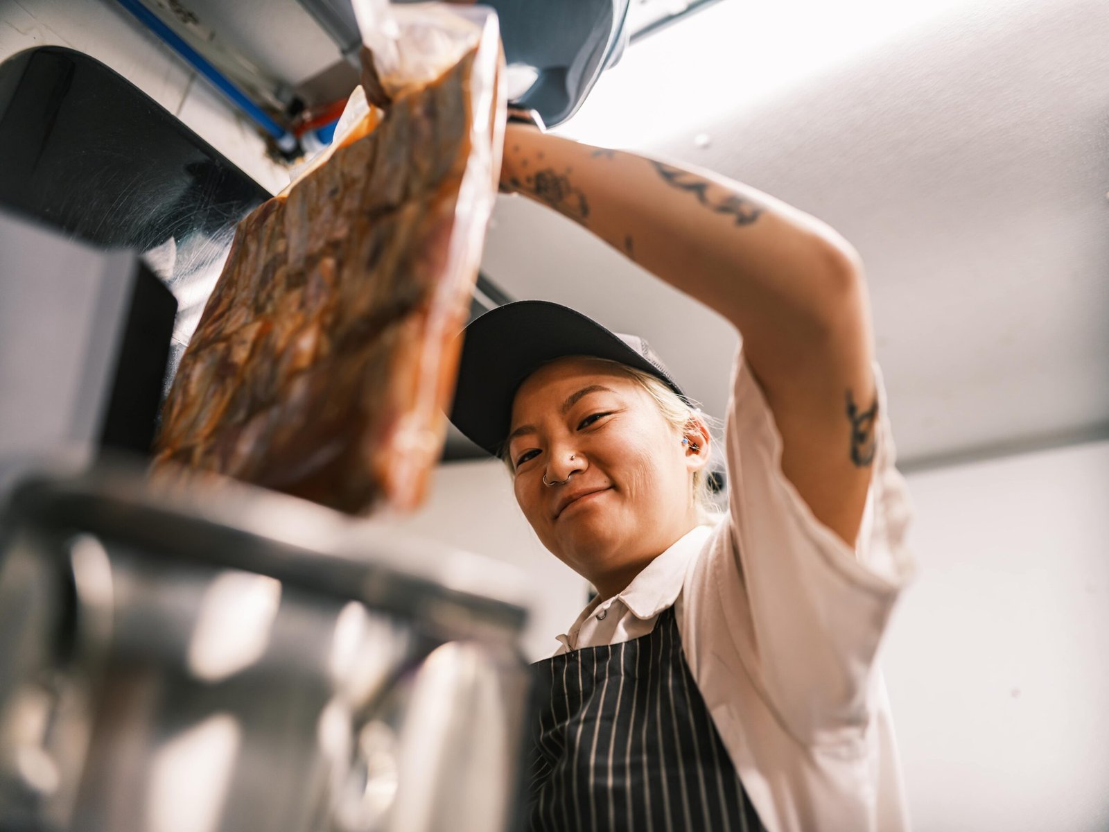 chef with sous vide pouch-min A chef wearing a striped apron and cap is placing a large vacuum-sealed pouch of food into a stainless steel container, likely preparing for sous vide cooking in a commercial kitchen setting. The chef's arms feature visible tattoos.