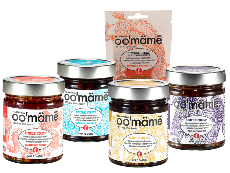 A selection of Oomame brand Chile Crisp products is displayed, featuring four glass jars with silver lids and colorful labels representing different global flavors: Szechuan, China (red label), Oaxaca, Mexico (blue label), Kerala India (yellow label), and Fez, Morocco (purple label). Behind the jars, there is a packet of Oomame Umami Heat Seasoning Blend (Japanese Togarashi) with an orange and beige design. The products are arranged on a white background.