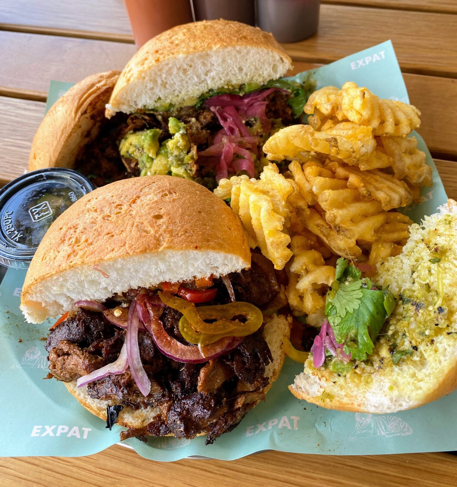 expat BBQ A close-up image of a plant-based sandwich meal featuring Harvest Shreds meat alternative. The sandwich is served on a soft bun and filled with seasoned Harvest Shreds, pickled onions, sliced jalapeños, guacamole, and fresh herbs. On the side are crispy waffle fries. Three bottles of sauce labeled "Awaze," "Baijiu," and "Hogwash" are visible in the background. The meal is presented on a tray lined with paper branded "EXPAT."