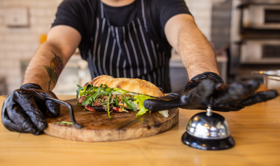 A chef wearing a black apron and black gloves presents a sandwich made with Harvest Shreds meat alternative and fresh greens on a wooden board, while pressing a service bell on a counter.