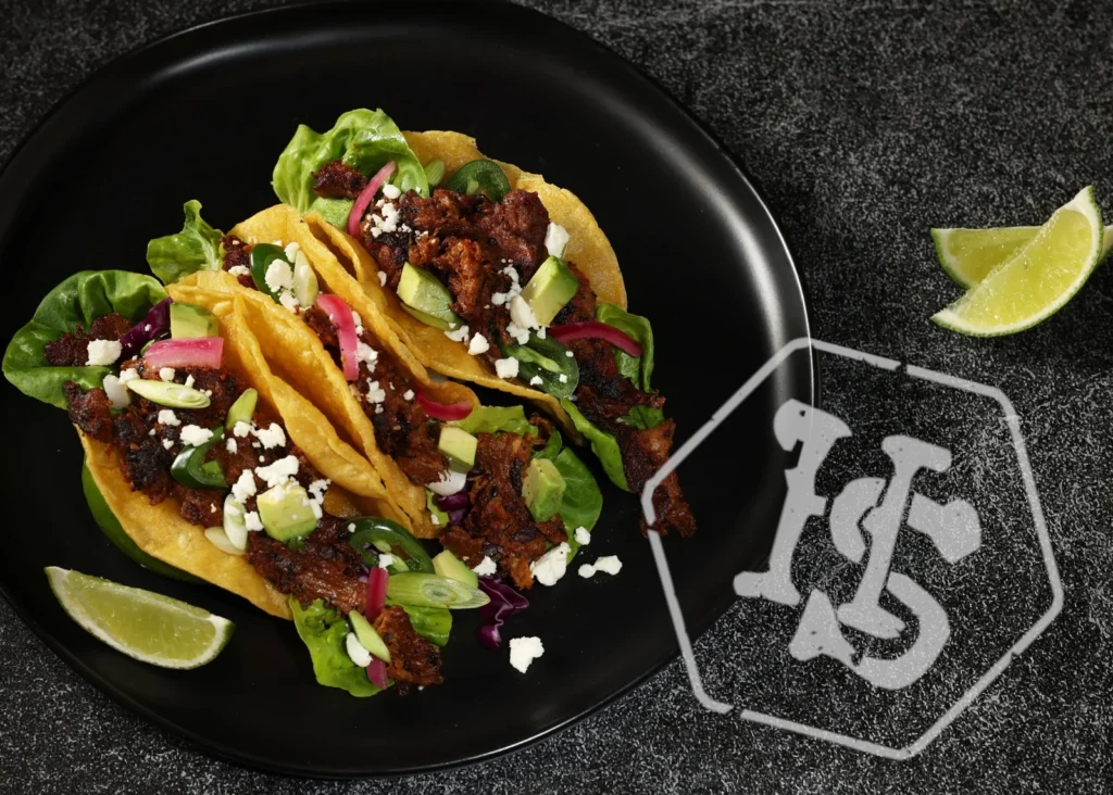 Harvest Shreds Tacos Harvest Shreds spiced with Barbacoa served in three corn taco shells with lettuce, pickled red onions, avocado chunks and queso fresco. On a black plate with lime garnish.